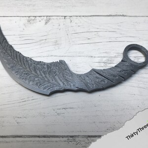Puede incluir: Un cuchillo de lanzamiento curvo de color plateado con un patr&oacute;n decorativo en la hoja. El cuchillo tiene un anillo en el mango.