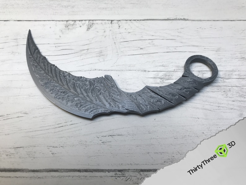 Puede incluir: Un cuchillo de lanzamiento curvo de color plateado con un patr&oacute;n decorativo en la hoja. El cuchillo tiene un anillo en el mango.