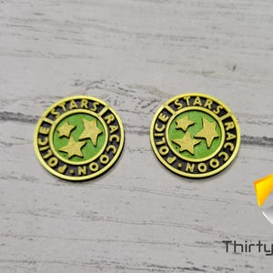 Puede incluir: Dos fichas de metal de color dorado con las palabras "STARS" y "POLICE" en un c&iacute;rculo alrededor de tres estrellas verdes. Las palabras "RACCOON" tambi&eacute;n est&aacute;n en el c&iacute;rculo.