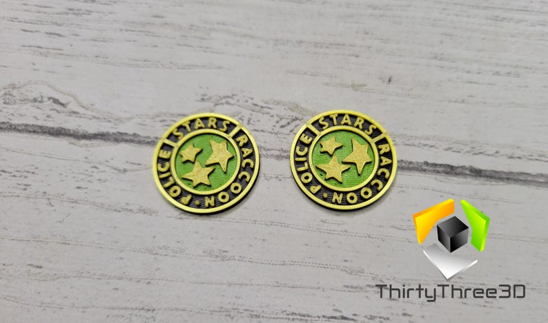 Puede incluir: Dos fichas de metal de color dorado con las palabras "STARS" y "POLICE" en un c&iacute;rculo alrededor de tres estrellas verdes. Las palabras "RACCOON" tambi&eacute;n est&aacute;n en el c&iacute;rculo.