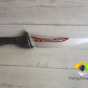 May include: A silver and red fantasy-style dagger with a black handle. The blade has a red design on it. The dagger is on a white wooden surface.
