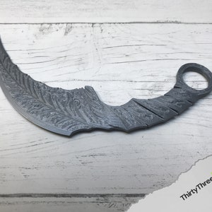 May include: A silver-colored, curved throwing knife with a decorative pattern on the blade. The knife has a ring on the handle.