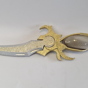 May include: An ornate dagger with a silver blade and gold-colored handle. The blade has a detailed pattern, and the handle features a bulbous, clear section and a red end. The design is intricate and decorative.
