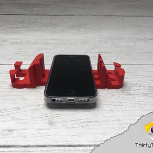 May include: A red 3D printed phone stand with a black smartphone resting on top. The stand is shaped like the word "GLASS".