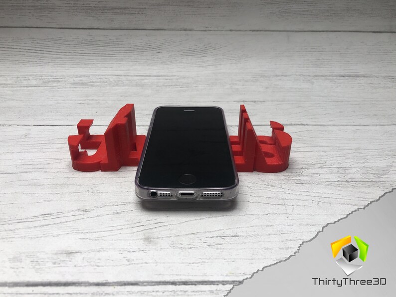 May include: A red 3D printed phone stand with a black smartphone resting on top. The stand is shaped like the word "GLASS".