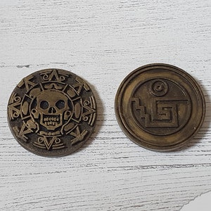 Puede incluir: Dos monedas de metal de color dorado con dise&ntilde;os intrincados. Una moneda presenta una calavera y huesos cruzados, mientras que la otra tiene un patr&oacute;n geom&eacute;trico.
