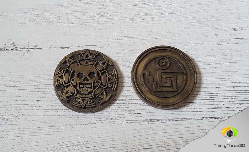 Puede incluir: Dos monedas de metal de color dorado con dise&ntilde;os intrincados. Una moneda presenta una calavera y huesos cruzados, mientras que la otra tiene un patr&oacute;n geom&eacute;trico.