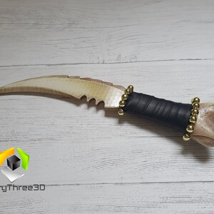 May include: A bone-like dagger with a curved blade and a serrated edge. The handle is wrapped in black leather and has gold beads around it.