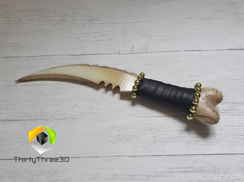 May include: A bone-like dagger with a curved blade and a serrated edge. The handle is wrapped in black leather and has gold beads around it.