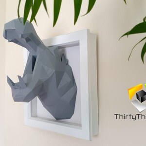 May include: A gray, geometric, 3D printed hippopotamus head mounted in a white frame. The hippopotamus has an open mouth and is facing the right side of the image. The frame is on a light beige wall. The text "ThirtyThree3D" is visible in the lower right corner of the image.