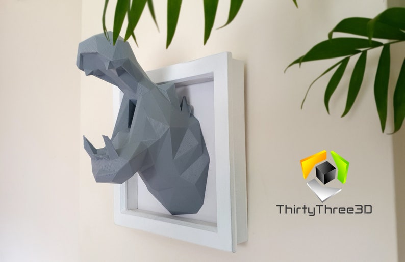 May include: A gray, geometric, 3D printed hippopotamus head mounted in a white frame. The hippopotamus has an open mouth and is facing the right side of the image. The frame is on a light beige wall. The text "ThirtyThree3D" is visible in the lower right corner of the image.