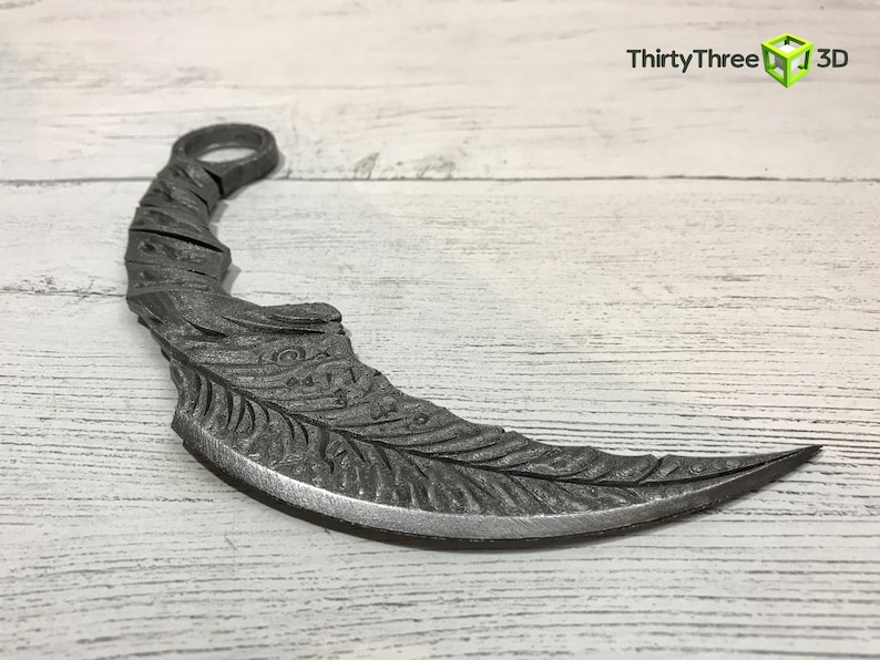 Puede incluir: Un cuchillo de lanzamiento de metal plateado con una hoja curva y un dise&ntilde;o de pluma detallado. El cuchillo tiene un anillo en la parte superior para sujetarlo.
