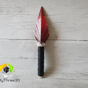 May include: A decorative spear with a red, leaf-shaped blade and a black handle wrapped in leather. The blade has a textured surface and is attached to the handle with white string. The spear is displayed against a white wooden background.
