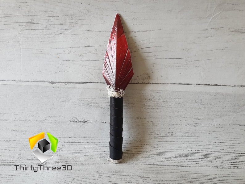 May include: A decorative spear with a red, leaf-shaped blade and a black handle wrapped in leather. The blade has a textured surface and is attached to the handle with white string. The spear is displayed against a white wooden background.