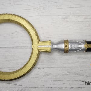 May include: A gold-colored, circular, decorative object with intricate markings. The object is attached to a silver and black handle.