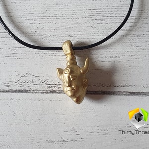 May include: Gold pendant necklace featuring a detailed sculpture of a demon's head with horns. The pendant is hanging from a black cord.