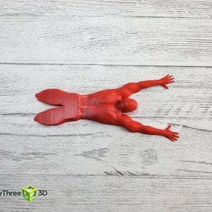 May include: Red 3D printed figurine of a person lying on their stomach with arms outstretched. The figurine is on a white wooden surface.