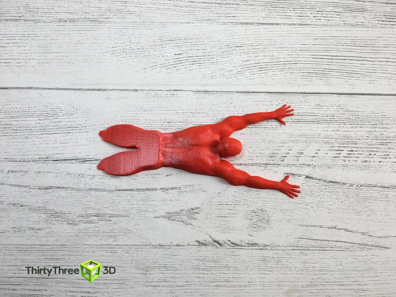 May include: Red 3D printed figurine of a person lying on their stomach with arms outstretched. The figurine is on a white wooden surface.