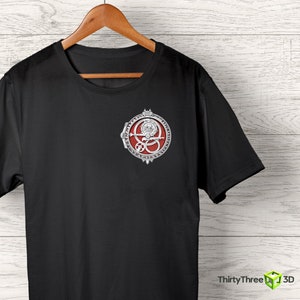 May include: Black t-shirt with a silver and red circular design featuring a symbol with a skull and crossbones. The symbol is surrounded by a circle of text.