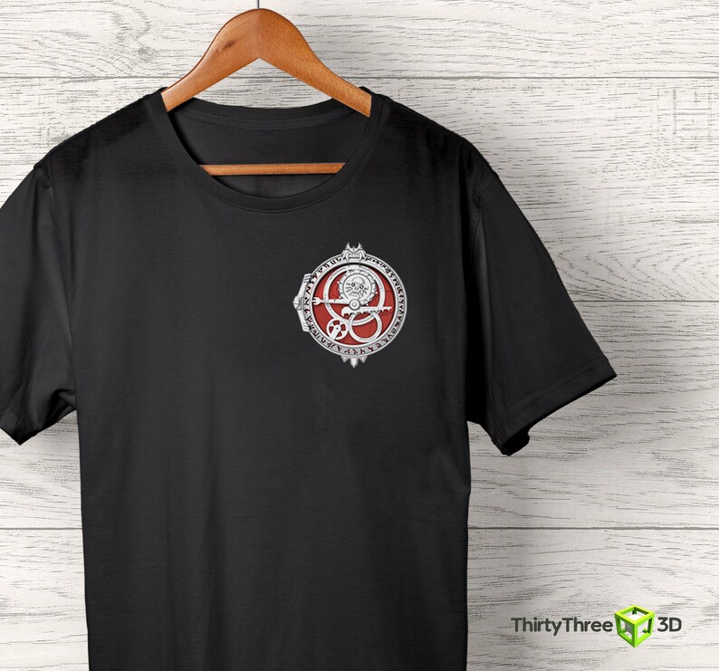 May include: Black t-shirt with a silver and red circular design featuring a symbol with a skull and crossbones. The symbol is surrounded by a circle of text.