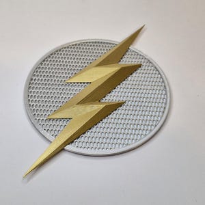 Puede incluir: Un emblema de rayo dorado, colocado sobre un fondo circular gris perforado. El emblema tiene una apariencia tridimensional, con &aacute;ngulos agudos y un brillo met&aacute;lico. El fondo tiene un borde blanco.