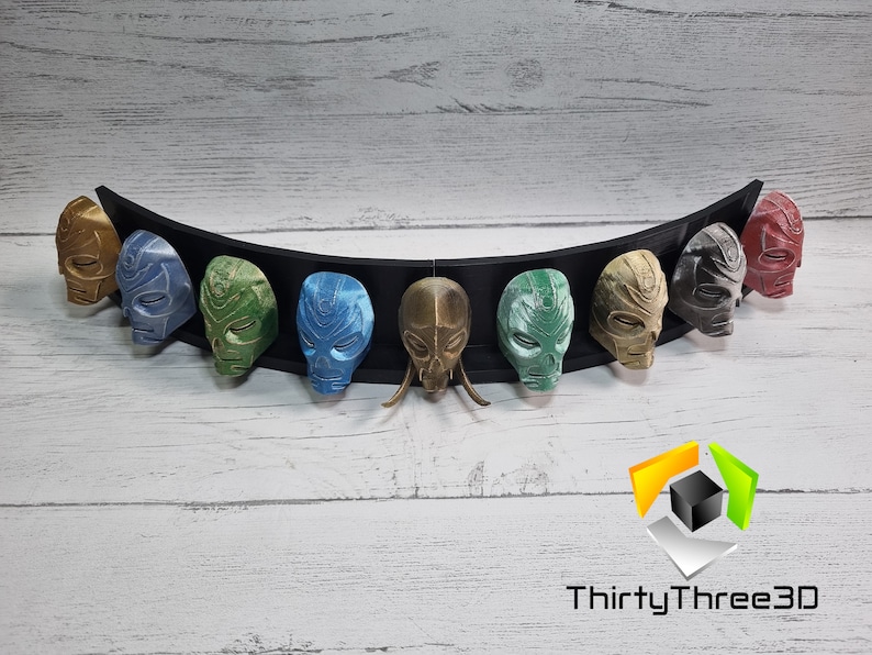 May include: A set of nine 3D printed masks, each painted in a different color, displayed on a black curved stand. The masks are all the same design, with a skull-like face and intricate details. The colors include gold, blue, green, brown, and red.