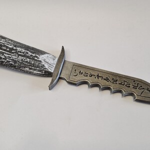 May include: A decorative dagger with a textured, gray and white handle. The blade is gray with a serrated edge and etched text. The dagger is a prop or collectible item.