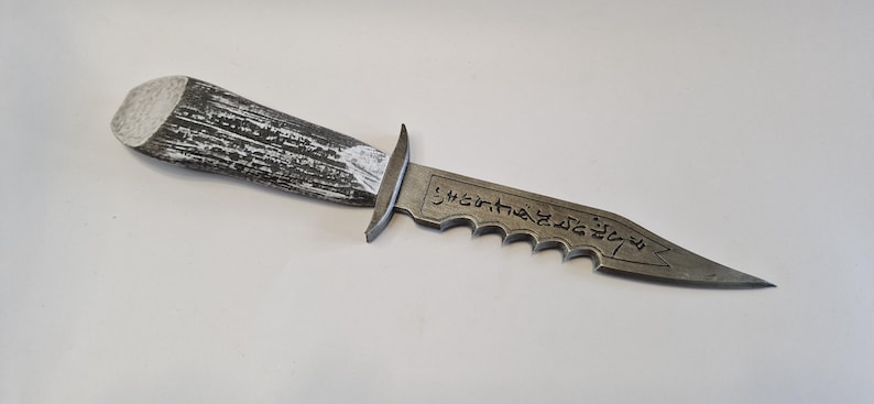 May include: A decorative dagger with a textured, gray and white handle. The blade is gray with a serrated edge and etched text. The dagger is a prop or collectible item.
