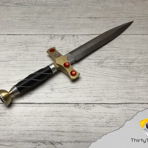 May include: A gold and black dagger with a silver pommel and red gems on the hilt. The dagger is lying on a white wooden surface.
