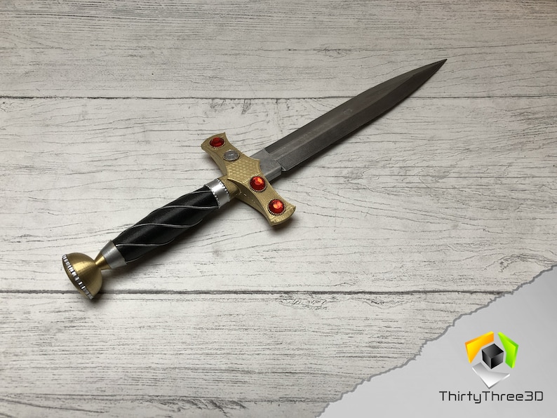 May include: A gold and black dagger with a silver pommel and red gems on the hilt. The dagger is lying on a white wooden surface.