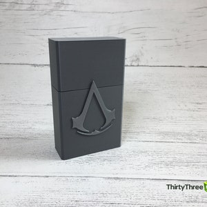May include: A gray rectangular box with a raised, silver-colored Assassin's Creed logo on the front.