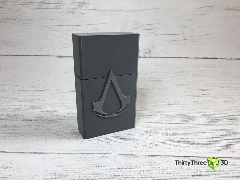 May include: A gray rectangular box with a raised, silver-colored Assassin's Creed logo on the front.