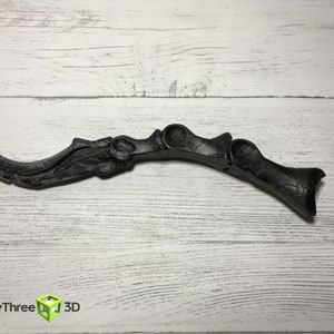 May include: A black, curved decorative object with a textured surface. The object has a blade-like shape with a series of rounded openings and a handle-like end. The object is set against a white wooden background.