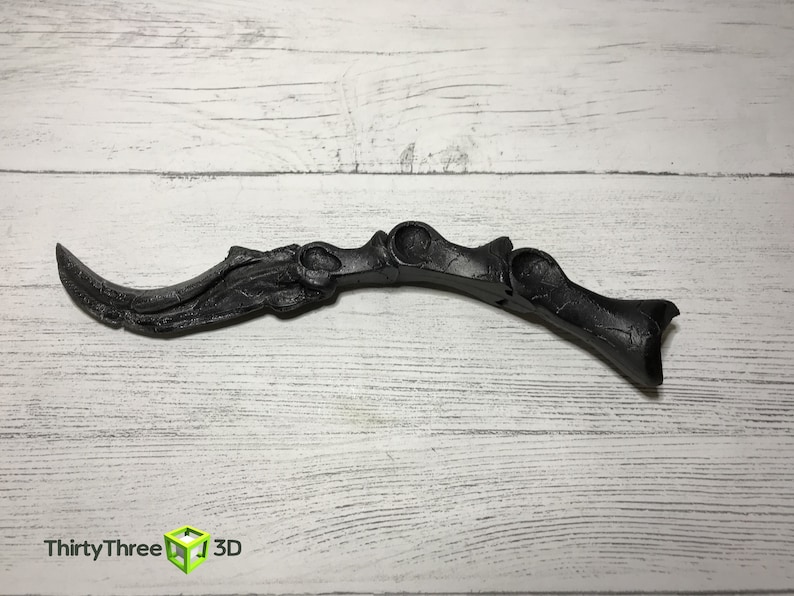 May include: A black, curved decorative object with a textured surface. The object has a blade-like shape with a series of rounded openings and a handle-like end. The object is set against a white wooden background.