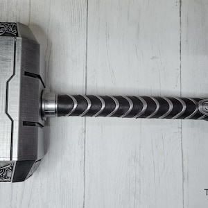 May include: A silver-colored Mjolnir hammer replica with a black leather handle and a strap. The hammer head has intricate details and a textured surface.