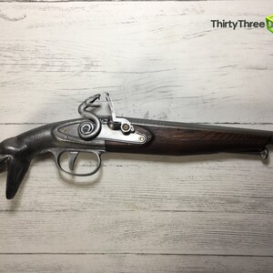 Puede incluir: R&eacute;plica de pistola de chispa con culata de madera oscura y acabado gris met&aacute;lico. La pistola presenta un dise&ntilde;o &uacute;nico con empu&ntilde;adura en forma de serpiente y elementos decorativos. El ca&ntilde;&oacute;n tiene una punta naranja.