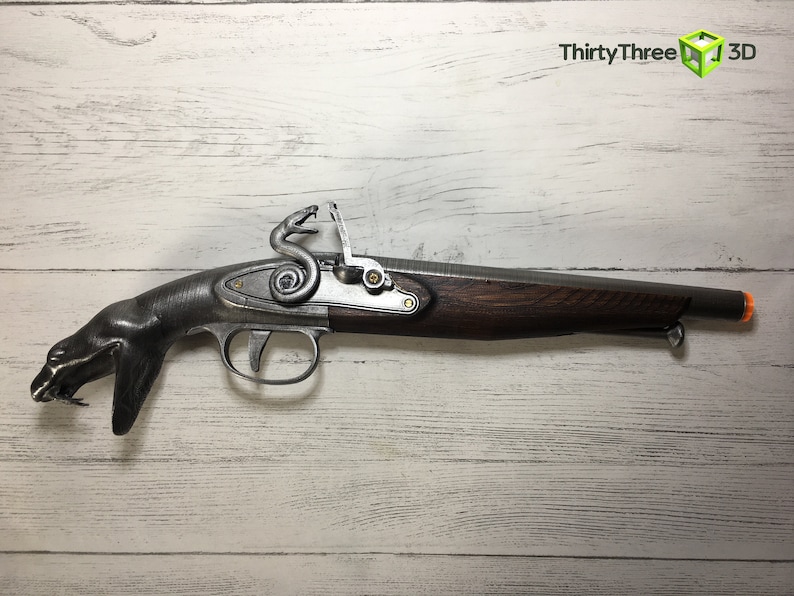 Puede incluir: R&eacute;plica de pistola de chispa con culata de madera oscura y acabado gris met&aacute;lico. La pistola presenta un dise&ntilde;o &uacute;nico con empu&ntilde;adura en forma de serpiente y elementos decorativos. El ca&ntilde;&oacute;n tiene una punta naranja.