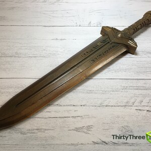 May include: A gold-colored sword with a detailed hilt and a blade inscribed with runes. The sword is lying on a white wooden surface.