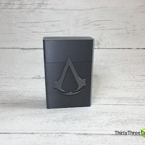 May include: A gray plastic box with a raised, black Assassin's Creed logo on the front.