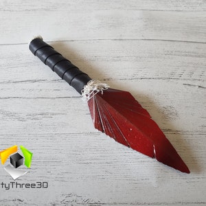 May include: A handcrafted decorative dagger with a red blade and a black leather-wrapped handle. The blade is shaped like a feather. The handle is wrapped in black leather and white string. The dagger is displayed on a light wood surface.