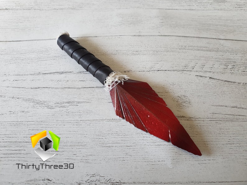 May include: A handcrafted decorative dagger with a red blade and a black leather-wrapped handle. The blade is shaped like a feather. The handle is wrapped in black leather and white string. The dagger is displayed on a light wood surface.
