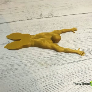 May include: A yellow 3D printed figurine of a person lying on their stomach with their arms outstretched. The figurine is on a white wooden surface.
