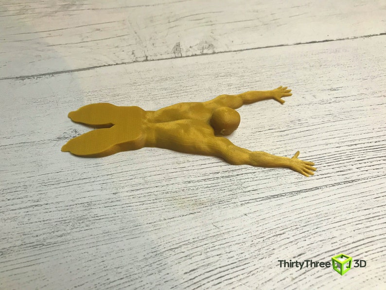 May include: A yellow 3D printed figurine of a person lying on their stomach with their arms outstretched. The figurine is on a white wooden surface.