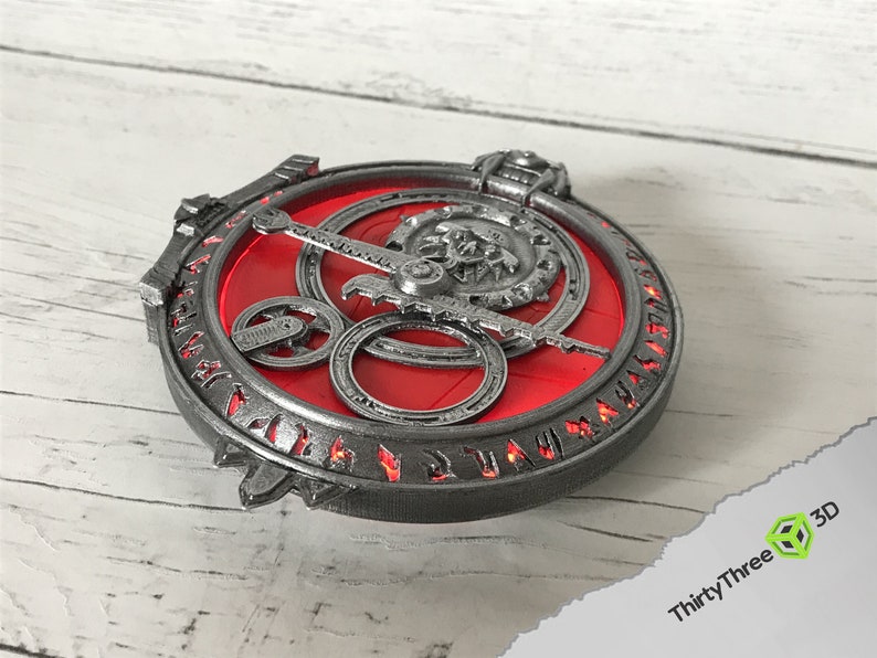 May include: A silver-colored metal pendant with a red center and intricate details. The pendant features a circular design with a central gear and surrounding gears. The outer edge of the pendant is inscribed with runes.