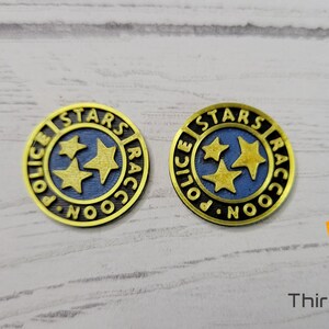 Puede incluir: Dos pines de metal de color dorado con las palabras "Stars Raccoon Police" y tres estrellas doradas sobre fondo azul.