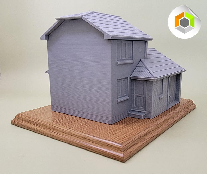 May include: A gray 3D printed model of a house with a wooden base. The house has a pitched roof, two windows, and a door.
