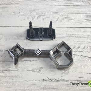 May include: A silver-colored metal key with a geometric design. The key is on a white wooden surface with a matching stand.