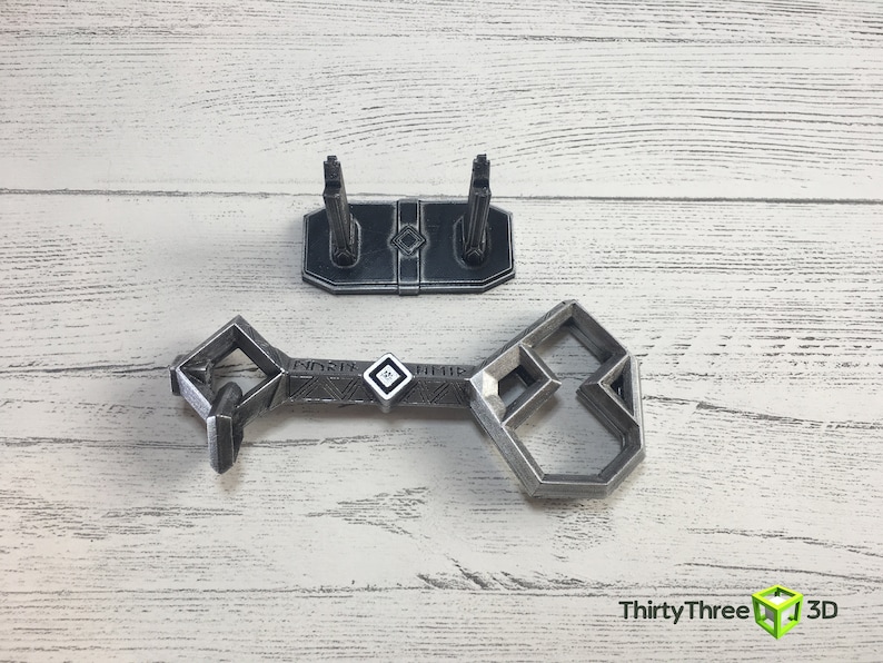 May include: A silver-colored metal key with a geometric design. The key is on a white wooden surface with a matching stand.