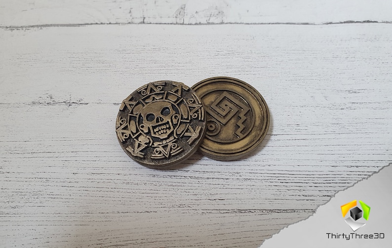 Puede incluir: Dos monedas de metal de color dorado con dise&ntilde;os intrincados. Una moneda presenta una calavera y huesos cruzados, mientras que la otra tiene un patr&oacute;n geom&eacute;trico.