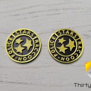 Puede incluir: Dos fichas de metal de color dorado con centros negros. Las fichas presentan las palabras "Raccoon City Police Stars" en un c&iacute;rculo alrededor de tres estrellas doradas.